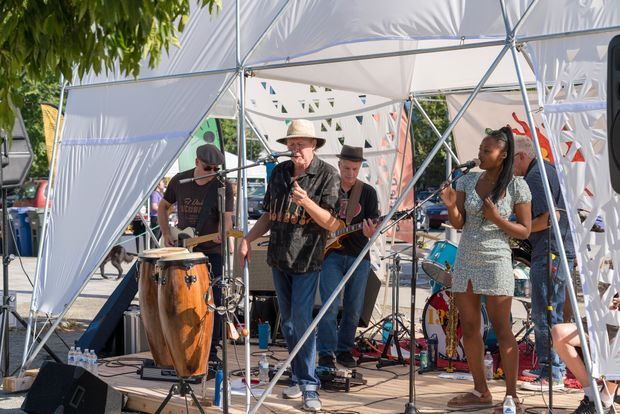 Outdoor live music performance on a small temporary stage, with several musicians, microphones, speakers, percussion instruments, guitars, and a geometric white fabric canopy structure. The image is set in a sunny outdoor event environment with trees and partial event infrastructure visible in the background.