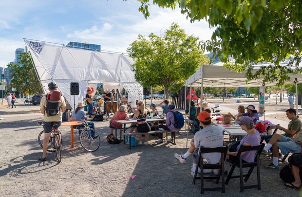 A sunny outdoor urban event space with people seated at picnic tables beneath a temporary canopy. A striking white geometric tensile pavilion and modern city buildings are visible in the background, with bicycles, trees, and hardscaped ground surrounding the gathering.