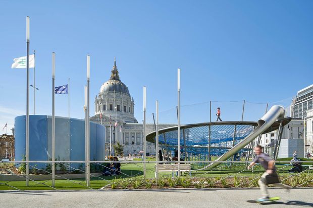 Bright daytime exterior view of a contemporary public plaza or streetscape with landscaped planting, a blue cylindrical structure, numerous flagpoles, and a distinctive elevated mesh pedestrian or recreational structure. A historic domed civic building is visible in the background, with people using the space and a modern building at right.