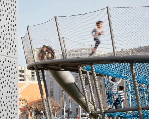 Outdoor elevated children's play structure in an urban setting, featuring a netted climbing and running platform, tubular metal supports, a large enclosed slide, and children using the equipment. A perforated architectural wall and surrounding mid-rise buildings are visible in the background.