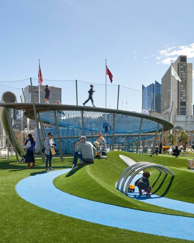 A vibrant urban outdoor recreation area with bright green artificial turf and winding blue pathways. The landscaped play space includes sculptural metal climbing arches, a large netted elevated pedestrian structure, flagpoles, and people using the park. High-rise and institutional-looking buildings form the city backdrop under a clear blue sky.