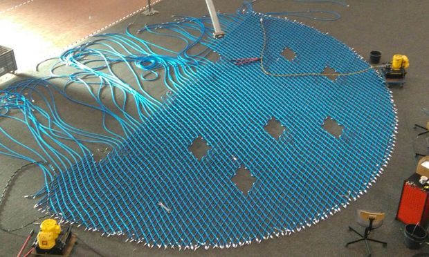 Overhead view of a large blue tubular mesh or hose assembly laid out on an industrial workshop floor, with numerous loose blue cables or tubes extending from one side. Small tools, equipment, and work stools surround the installation. The image documents a technical fabrication or construction process rather than a finished room or building.
