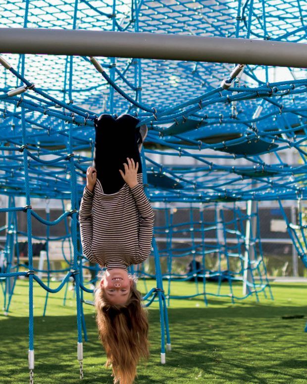 A child hangs upside down from a blue rope climbing structure in an outdoor playground. The image is dominated by a large blue netted play frame over artificial grass, with surrounding playground elements and a building visible in the background.