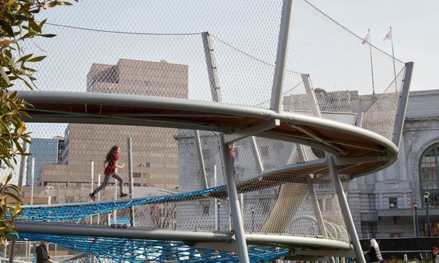 An elevated pedestrian walkway or bridge with sweeping curved metal structures and chain-link safety fencing, crossing above a lower level in an urban setting. People are walking along the structure, with large institutional or commercial buildings visible behind it.