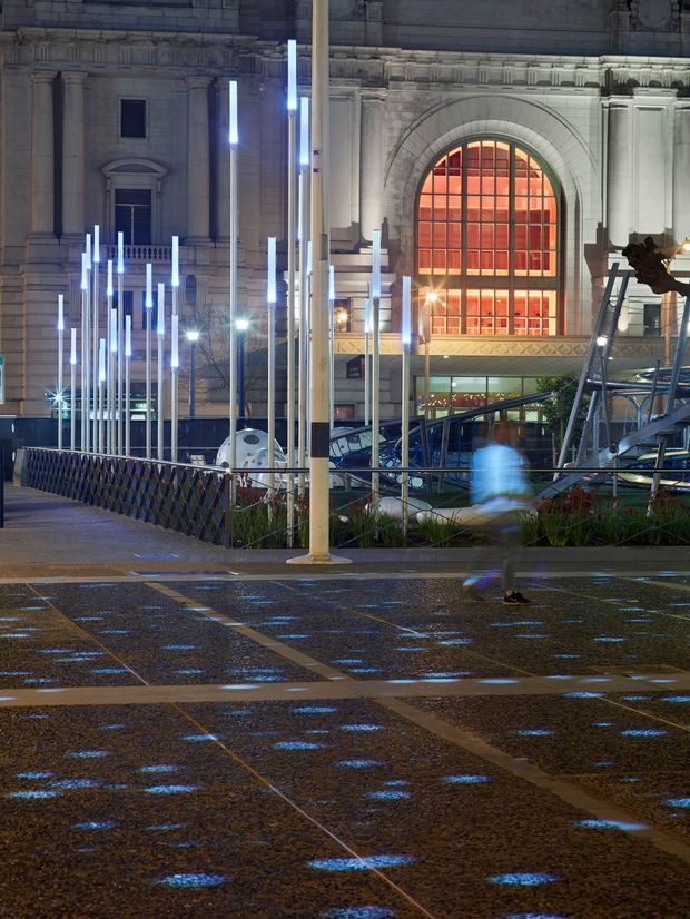Night-time view of a grand heritage-style urban building and public plaza. The stone facade features classical columns, ornate detailing, and a large arched window glowing orange from within. In the foreground, a contemporary public art installation of tall illuminated vertical poles stands beside a wet paved promenade with blue reflected light, railings, planted areas, and blurred pedestrians.