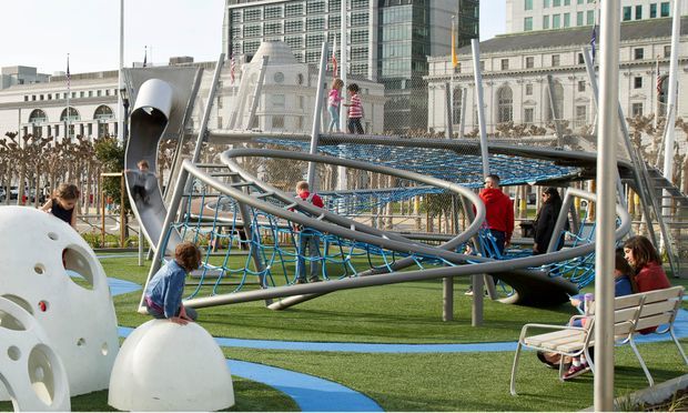 A contemporary urban playground with children climbing and playing on sculptural white climbing forms, rope-net structures, slides, and curved grey metal frames. The play area is surfaced with artificial green turf and bright blue pathways, with benches around the perimeter. Historic and modern civic buildings form the dense city backdrop.