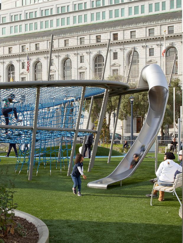 A landscaped urban playground with a large climbing net structure and a sculptural stainless-steel tube slide. The play area has artificial turf, curved concrete edging, trees, and seating, set against an ornate multi-storey civic or institutional building facade with arched windows, decorative stonework, and flags.