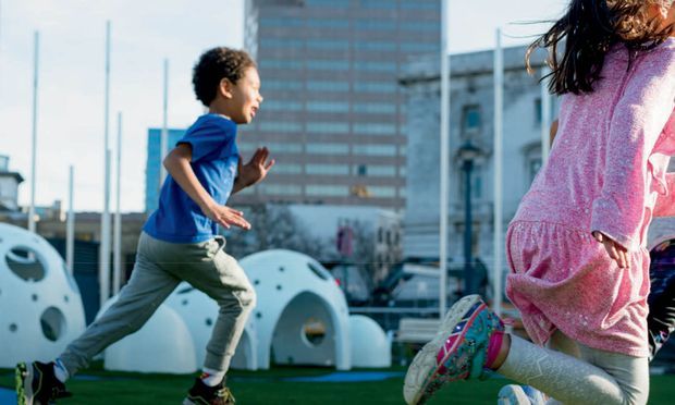 Outdoor urban scene with children running across a landscaped recreational area, featuring sculptural white structures and modern city buildings in the background.