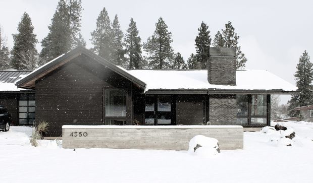 Snow-covered single-storey residential house in a wooded setting, with a dark timber-clad gabled wing, broad low-pitched roof, exposed brick chimney and large windows. A concrete entrance wall marked 4350 sits in the snowy foreground.