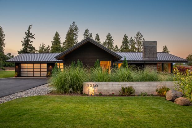 Single-storey contemporary residential home viewed from the front at dusk, set within a landscaped suburban or semi-rural garden. The house has dark horizontal cladding, broad low-pitched metal roofs, a prominent central gabled entry volume, warm timber-toned entry doors, extensive garage glazing, and a substantial brick chimney. Ornamental grasses, lawn, gravel edging, and a timber retaining planter wall frame the facade.