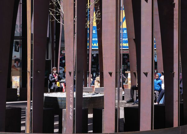 Urban commercial or civic plaza viewed through a series of tall rust-coloured steel fins. The vertical architectural screen creates a strong rhythmic facade, with reflective glazing, integrated bench seating, a shallow rectangular water feature or planter, small trees, and people visible in the public space beyond. The scene has a bold, contemporary and slightly industrial character with strong sunlight and high contrast.