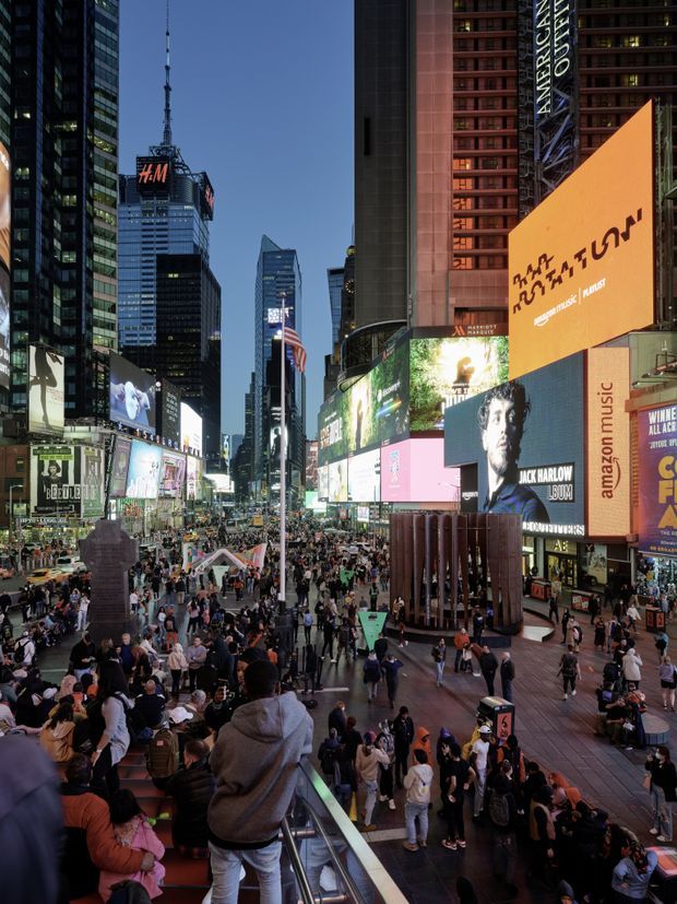 A dusk street-level view from elevated steps overlooking a dense urban entertainment district, dominated by towering commercial buildings, illuminated digital billboards, retail signage, crowds of pedestrians, and broad paved public plazas. The scene is strongly metropolitan and commercial, with high-rise architecture and intense advertising displays.