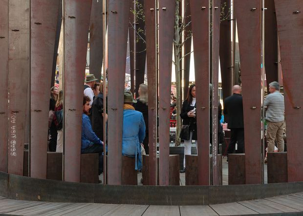 An outdoor public installation or architectural screen made from tall, irregularly spaced weathered steel plates, with visible bolts and narrow openings. People and small trees are seen behind the sculptural metal enclosure, while the foreground has a curved dark metal base and paved decking. The composition feels industrial and urban, with the rust-toned steel structure dominating the frame.