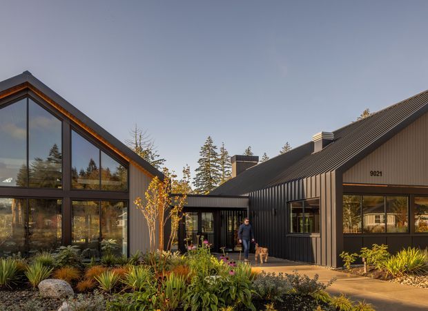 Exterior view of a modern, dark-clad residential building composed of connected gabled volumes. The left volume has expansive floor-to-ceiling glazing, while the central and right sections feature vertical dark cladding, pitched roofs, and large windows. Native-style landscaped planting and a paved entry path occupy the foreground.