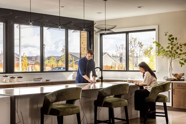 A bright contemporary kitchen with a large island, integrated sink and tap, pale veined stone surfaces, dark-framed windows, and three upholstered bar stools. Two people are using the island, while pendant lights hang above. A timber-toned built-in cabinet and indoor plant add warmth to the neutral space.