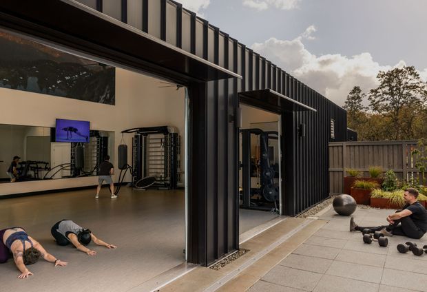 Contemporary black-clad building with large open glazed doors leading to a spacious home gym. The gym contains exercise equipment, wall mirrors, a television, and people exercising, while the adjoining paved outdoor area has free weights, a large dark spherical feature, timber fencing, and planted containers.