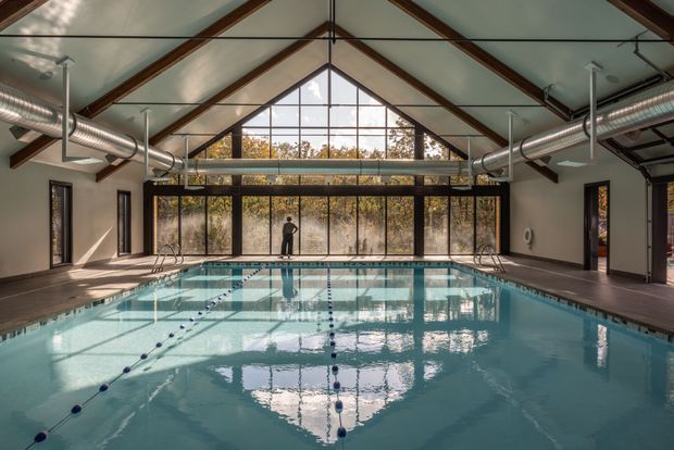 A large indoor swimming pool housed in a pavilion-like building with a steep gabled glazed roof and full-height windows. Exposed timber roof beams, dark-framed glazing, polished concrete or tiled pool surrounds, and prominent exposed silver ventilation ducts create a contemporary recreational space. Lane markers and pool equipment are visible, with strong reflections across the water.