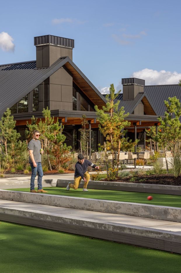 Exterior view of a contemporary lodge-style building with steep dark metal gabled roofs, tall capped chimneys, dark cladding, and warm timber soffits. The building opens onto a landscaped outdoor recreational area with synthetic lawn bowling or bocce-style lanes, planting, seating, and people using the space.
