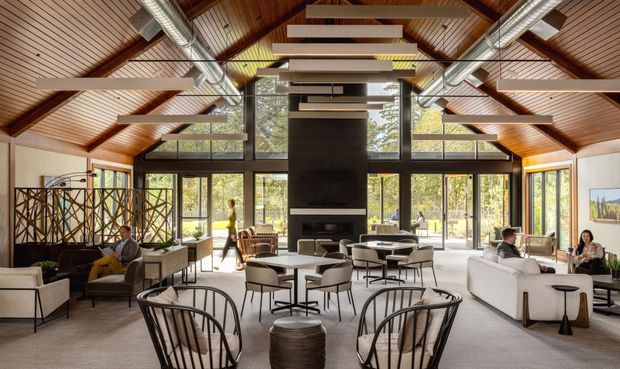 A spacious, light-filled living room with a dramatic high pitched timber-lined ceiling, exposed dark timber beams, suspended linear feature lighting, and exposed metal ductwork. Full-height black-framed glazing surrounds the room and opens to the outdoors. The space is arranged with multiple lounge seating groups, armchairs, coffee tables, and a central fireplace and television wall. Decorative timber screens divide a seating area on the left, while contemporary furnishings create a refined lodge-like atmosphere.