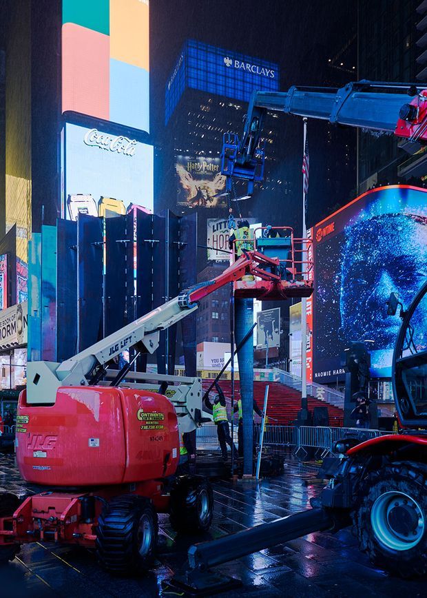A nighttime urban streetscape crowded with illuminated digital billboards, including Barclays, Coca-Cola and Harry Potter signage. Construction lifts and equipment occupy the wet pavement in the foreground, with intense blue, pink and red lighting reflecting across the scene. The image is primarily a commercial city exterior rather than an architectural interior.