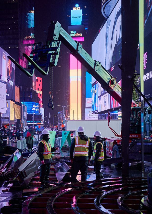 A nighttime urban construction scene in a brightly illuminated city plaza, with construction workers in high-visibility vests and hard hats standing beside a large crane and truck. Wet pavement reflects vivid pink, blue, and white digital billboards, while metal construction components and barriers occupy the foreground. The image documents active city-center infrastructure or building work rather than an architectural interior.