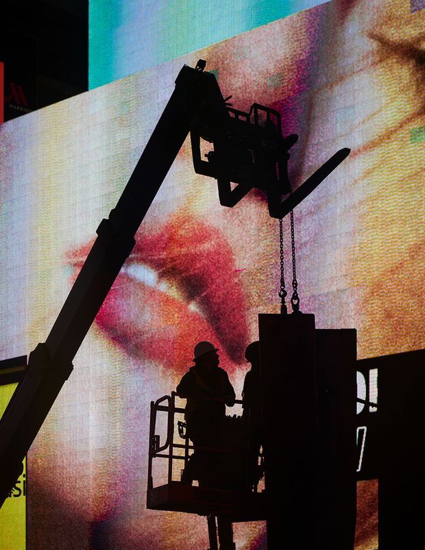 A dramatic silhouetted construction or production scene, with two workers standing in an elevated basket beside a large articulated boom lift or crane. Chains suspend a bulky piece of equipment, while a giant colorful LED screen or illuminated backdrop fills the background. The image is high-contrast and industrial, with strong red, orange, blue, and black tones.