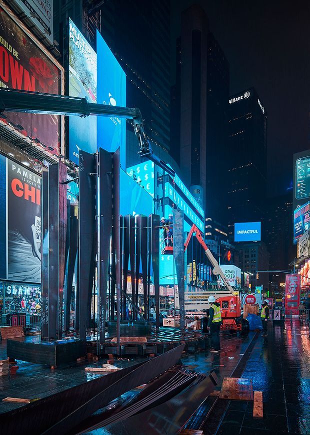 Nighttime urban streetscape in a dense commercial entertainment district, with towering buildings covered in illuminated digital billboards, wet reflective pavement, construction equipment, workers, and temporary street structures. The scene strongly resembles Times Square or a comparable major city advertising plaza.