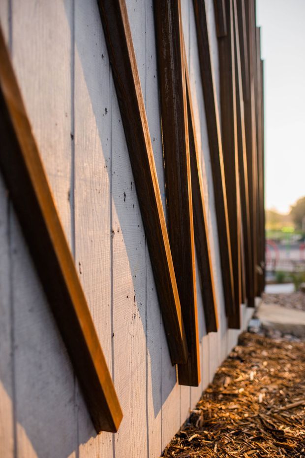 Close-up exterior architectural detail of a wall clad in pale timber boards, with repeated dark timber vertical and angled battens creating a strong rhythmic facade pattern. Warm low-angle sunlight emphasizes the wood grain and shadows; landscaped ground and a blurred residential setting are visible beyond.