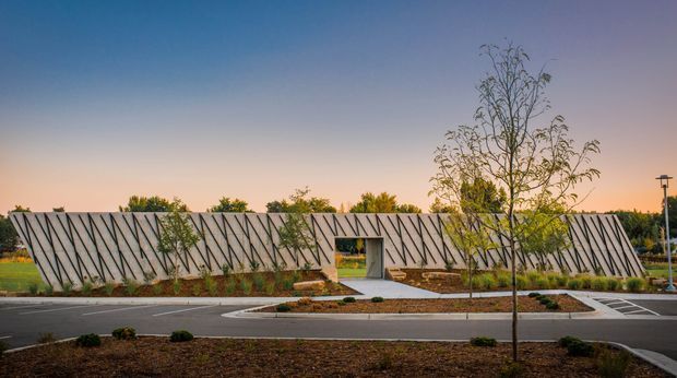 Wide exterior view of a low, contemporary architectural structure set within a landscaped site at dusk. The long pale wall is patterned with repeated dark diagonal slats, creating a strong geometric facade, with a central recessed entrance and paved path. Newly planted trees, garden beds, curving roadways, and clean site landscaping surround the building.