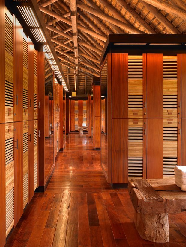 A warm, richly timber-lined interior corridor with extensive built-in locker or wardrobe-style cabinetry on both sides. The space has a polished timber floor, slatted timber cabinet fronts, dark trim, and a high exposed timber post-and-beam roof structure. A rustic timber bench with folded towels occupies the foreground, suggesting a resort changing area, spa, or sauna-adjacent facility.