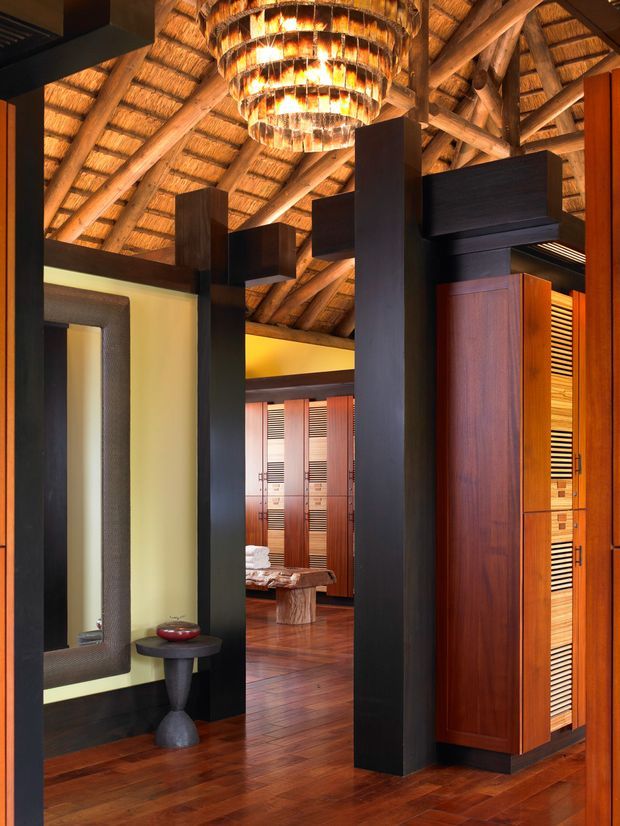 A richly detailed interior entry or circulation space beneath a dramatic timber thatched roof. Tall dark timber architectural screens and posts frame views into a warm wood-lined room, with built-in timber cabinetry, polished timber flooring, a sculptural side table, and a large layered pendant light. The palette is warm, earthy, and luxurious, with strong contrast between dark-stained timber and golden natural wood.