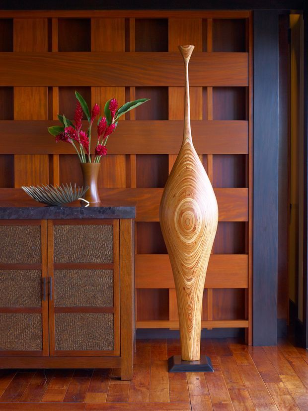 A warmly lit interior vignette featuring rich timber wall panelling with a geometric grid of recessed sections, timber flooring, and a dark stone-topped woven-front cabinet. A tall sculptural carved timber object stands on a plinth beside a vase of red flowers and a decorative metal bowl. The composition has a strong mid-century-inspired, handcrafted character.