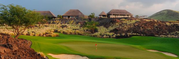 A landscaped golf course with a manicured green, sand bunkers, rugged dark volcanic terrain, and several low-rise buildings with hipped, thatched-looking roofs on the hillside in the background.