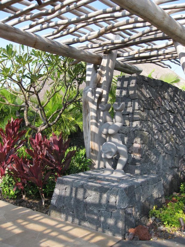 A tropical outdoor garden alcove with a rustic timber pergola made from substantial natural branches and trunks. A dark volcanic-stone masonry bench and wall sit beneath the pergola, with a small sculptural figure on the bench. Lush tropical planting surrounds the structure, and strong sunlight creates striped shadows across the stone and paving.