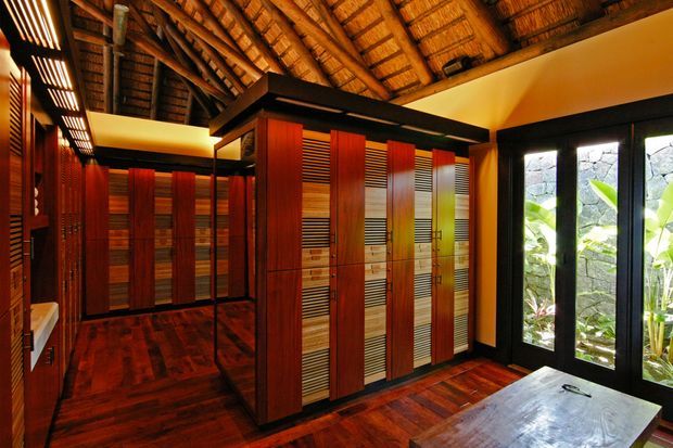 A richly detailed, warm-toned interior dominated by extensive built-in timber cabinetry and wardrobe storage. The room features deep reddish-brown timber flooring, vertical timber panels, louvred cabinet doors, black framing, and a dramatic exposed thatched or timber-lined pitched ceiling. Full-height glazed doors open views toward tropical planting and a stone wall outside.