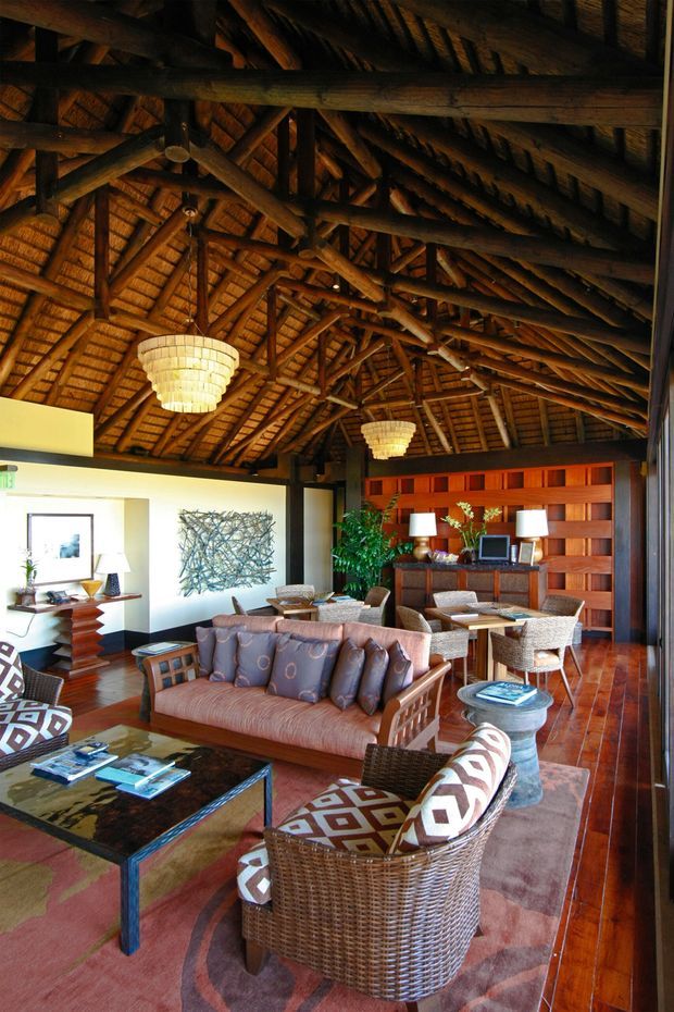 A warm, richly detailed living room beneath a dramatic vaulted timber roof with exposed rustic rafters and heavy post-and-beam construction. The room features woven rattan lounge chairs, a patterned upholstered sofa, a dark glass coffee table, timber flooring, a timber feature wall with built-in cabinetry, framed artwork, plants, and pendant lighting. The interior has a lodge-like, tropical-resort atmosphere with strong natural materials and high ceilings.