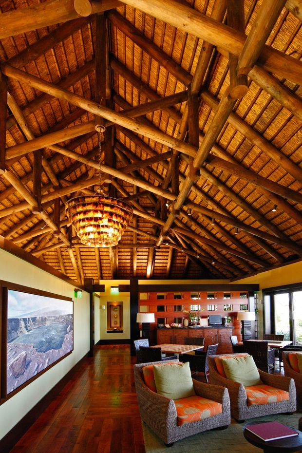 A richly detailed lodge-style living room beneath a dramatic high pitched roof with exposed round timber beams and a woven timber or thatched roof lining. The space includes dark timber flooring, woven lounge chairs with patterned orange cushions, a low coffee table, table lamps, a large wall-mounted artwork, and a timber bar or partition with open shelving. Warm amber lighting creates a rustic, luxurious atmosphere.