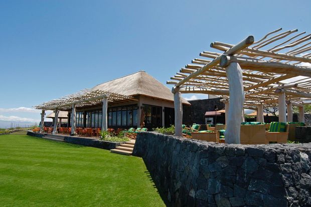 A low-rise resort-style building with a broad thatched roof, extensive timber pergola structures, dark volcanic-stone retaining walls, and outdoor dining/lounge areas overlooking an open landscape. The setting is sunny and tropical or coastal, with manicured lawn and strong indoor-outdoor connection.