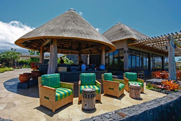 Open-air tropical resort-style outdoor lounge and bar with two large thatched pavilion roofs, timber posts, a pergola, stone paving, and colourful upholstered armchairs arranged around small drum tables. Lush planting and bright blue sky surround the relaxed entertaining area.