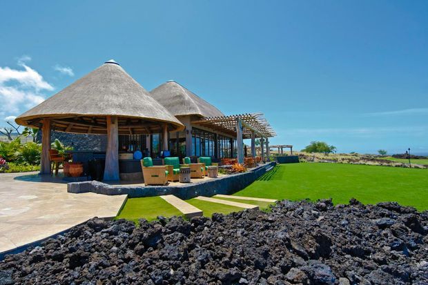 A tropical resort-style pavilion beside a broad manicured lawn, with multiple conical thatched roofs, open timber-framed dining and lounge areas, a pergola, paved terraces, and dark volcanic rock in the foreground. Ocean and bright blue sky are visible in the distance.