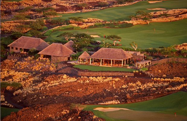 Aerial view of a low-rise resort-style complex set within a dramatic arid volcanic landscape and surrounded by manicured green golf-course fairways. Several pavilion-like buildings have broad hipped roofs, with a larger open-sided central building and landscaped outdoor areas.