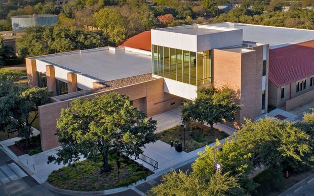 Aerial view of a substantial contemporary institutional or commercial building set within a leafy, landscaped urban campus. The complex combines warm red-brown brick walls, extensive glazing, crisp white rectangular volumes, flat roofs, and a prominent maroon pitched roof section. Paved paths, mature trees, planting beds, and accessible entry ramps surround the building.