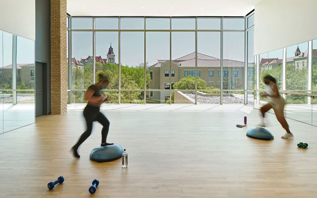A bright, spacious fitness studio with pale timber flooring, full-height glazed walls, mirrored side walls, and a warm exposed brick column. Two people are exercising with balance trainers and small hand weights. The glazing overlooks landscaped grounds and neighbouring buildings, creating an airy, open atmosphere.
