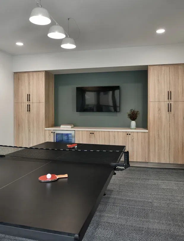 A contemporary recreation room with a full-size table tennis table in the foreground. The rear wall features light timber built-in cabinetry surrounding a dark green television feature wall, with a low storage console, wall-mounted TV, pendant lights, recessed ceiling lighting, and grey patterned carpet.