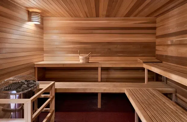 A compact sauna interior lined throughout with warm-toned horizontal timber boards and a slatted timber ceiling. Tiered built-in benches wrap around the room, with a sauna heater and stones in the foreground, a small bucket on the rear bench, and a warm wall light creating an intimate, enclosed atmosphere.
