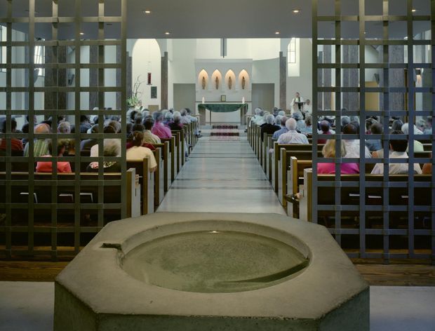 Interior view looking down the central aisle of a contemporary church or chapel during a service. Rows of timber pews are filled with congregants, framed by dark gridded screens, leading to a raised altar with a cross and sculptural wall niches. A large stone baptismal font dominates the foreground.