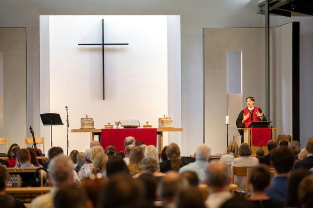 Interior view of a contemporary church sanctuary during a service, with a large illuminated cross on the rear wall, altar dressed in red, lectern, clergy member, musical instruments, and a seated congregation. The space has restrained neutral finishes, strong vertical and horizontal architectural elements, and a high, open ceiling area.