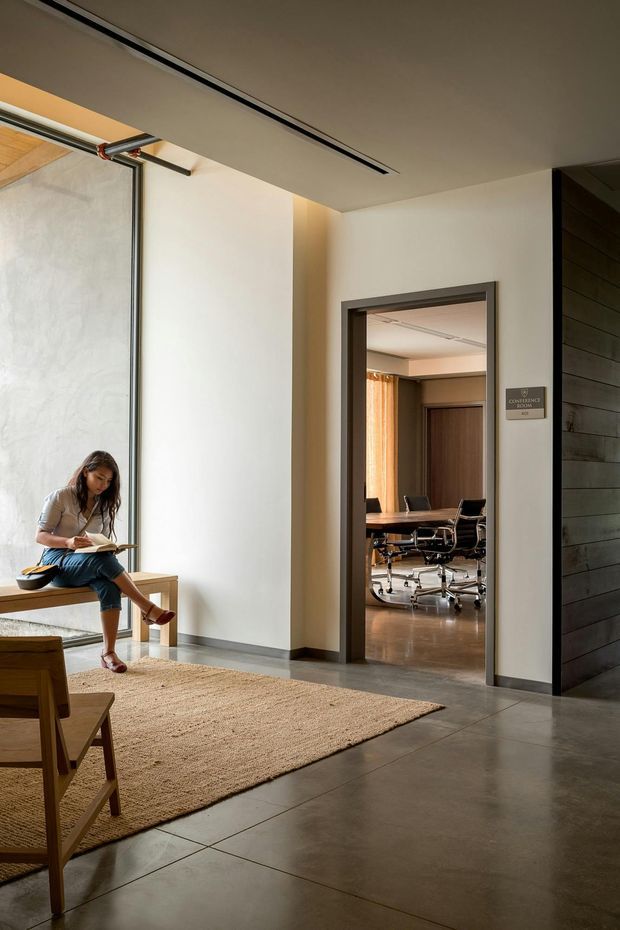 A refined contemporary interior showing a spacious lounge or waiting area opening into a conference room. The space features polished concrete or large-format grey flooring, warm off-white plaster walls, a neutral woven rug, timber seating, a full-height glazed sliding partition with exposed hardware, and dark timber wall cladding. A woman sits reading near the window, while the adjoining meeting room contains a conference table and rolling office chairs. Recessed linear ceiling lighting and warm daylight create a calm, professional atmosphere.