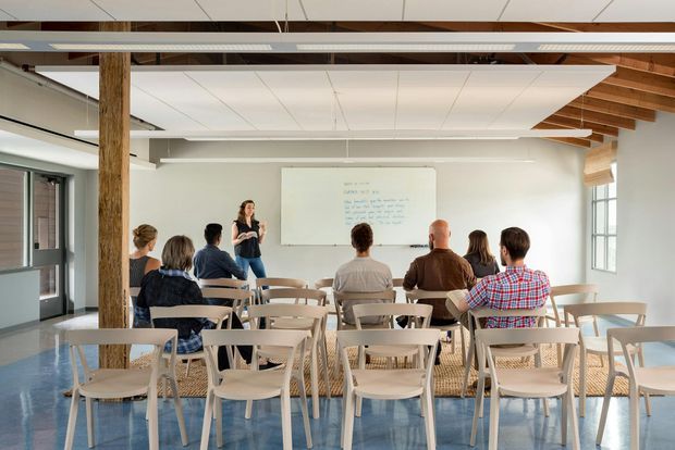 Bright open-plan teaching or meeting space with rows of light timber chairs and tables facing a presenter and projection screen. The room features exposed timber posts and roof rafters, a white suspended ceiling, large industrial-style windows, and a polished blue-grey floor.
