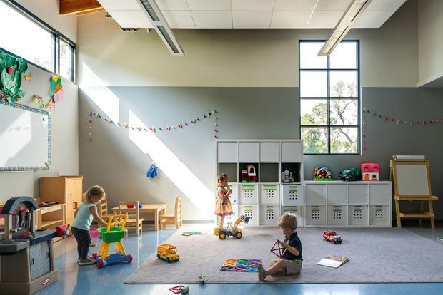 Bright, spacious children's playroom or early-learning room with large windows, high ceilings, a carpeted play area, low white storage cubbies, child-sized timber furniture, toys, and colourful bunting. The room has pale grey-green walls, a tiled suspended ceiling with exposed linear services, and strong natural sunlight.